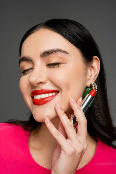 Retrato de mujer elegante con pendientes de moda, maquillaje impecable y cabello moreno sosteniendo lápiz labial rojo entre los dedos y sonriendo sobre fondo gris - foto de stock