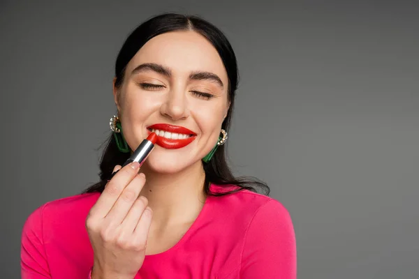 Porträt einer schicken jungen Frau mit trendigen Ohrringen und makellosem Make-up, die roten Lippenstift aufträgt und mit geschlossenen Augen lächelt, während sie auf grauem Hintergrund posiert — Stockfoto