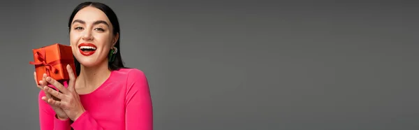 Animado e chique jovem mulher com brincos na moda e cabelo morena sorrindo enquanto segurando caixa de presente vermelha e embrulhada no fundo cinza, banner — Fotografia de Stock