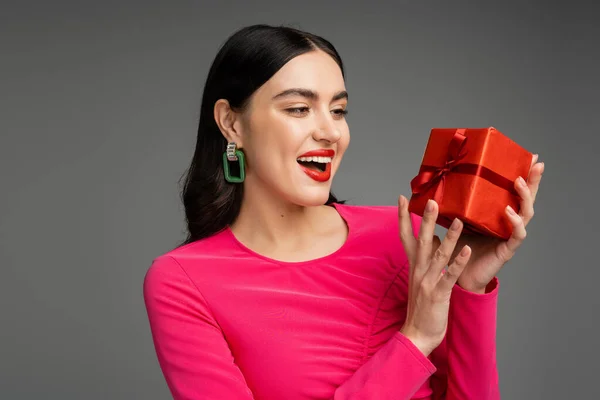Mujer joven excitada y elegante con pendientes de moda y pelo morena brillante sonriendo mientras sostiene la caja de regalo roja y envuelta en fondo gris - foto de stock