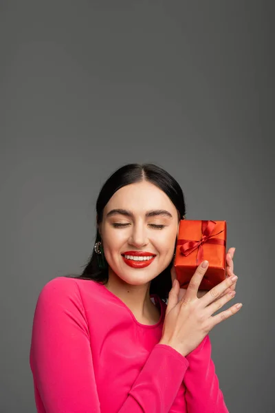 Verführerische und schicke junge Frau mit trendigen Ohrringen und glänzenden brünetten Haaren lächelt, während sie mit geschlossenen Augen posiert und eine rot verpackte Geschenkbox auf grauem Hintergrund hält — Stockfoto