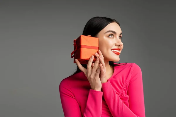 Чарівна і шикарна молода жінка з модними сережками і блискучою брюнеткою, що посміхається, тримаючи червону і загорнуту подарункову коробку на сірому фоні з місцем для тексту — стокове фото