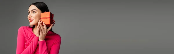 Bezaubernde und schicke junge Frau mit trendigen Ohrringen und glänzenden brünetten Haaren, die lächelt, während sie rote und umwickelte Geschenkschachteln auf grauem Hintergrund hält, Banner — Stockfoto
