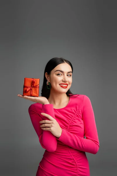 Seductora y feliz joven con pendientes de moda y pelo morena brillante sonriendo mientras sostiene el regalo rojo y envuelto en fondo gris - foto de stock