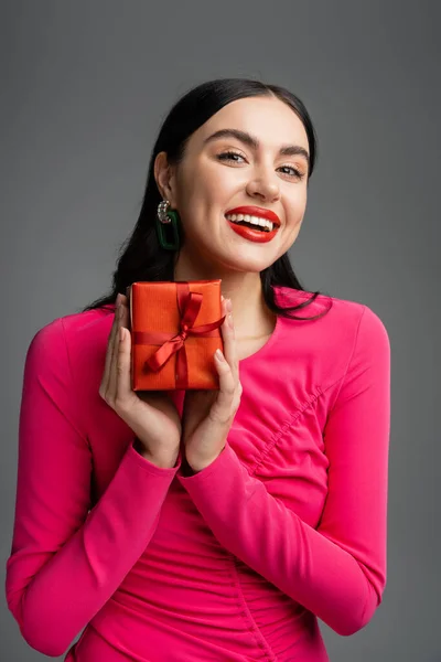 Positive and young woman with trendy earrings and brunette hair smiling while holding red and wrapped present for holiday on grey background — Stock Photo