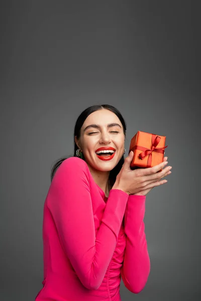 Aufgeregte junge Frau mit trendigen Ohrringen und brünetten Haaren, die lächelnd mit geöffnetem Mund steht und ein rot verpacktes Geschenk für den Urlaub auf grauem Hintergrund in der Hand hält — Stockfoto