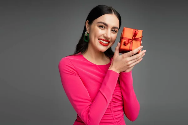 Mujer elegante y feliz con pendientes y cabello moreno sonriendo mientras sostiene el regalo rojo y envuelto para las vacaciones sobre fondo gris - foto de stock