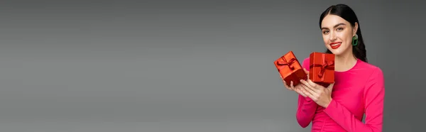 Glückliche Frau mit brünetten Haaren, die lächelnd im magentafarbenen Partykleid steht und verpackte und rote Geschenkboxen für den Urlaub auf grauem Hintergrund hält, Banner — Stockfoto