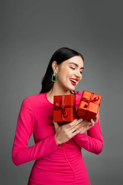Веселая молодая женщина с блестящими брюнетками и модными серёжками, улыбающаяся стоя в пурпурном платье для вечеринок и держа упакованные подарочные коробки для отдыха на сером фоне — стоковое фото