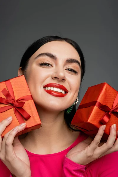 Portrait of cheerful young woman with shiny brunette hair and trendy earrings smiling while holding wrapped gift boxes for holiday and looking at camera on grey background — Stock Photo