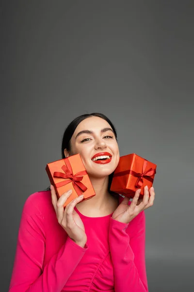 Verführerische junge Frau mit glänzenden brünetten Haaren lächelt, während sie im magentafarbenen Partykleid steht und verpackte Geschenkboxen für den Urlaub auf grauem Hintergrund hält — Stockfoto