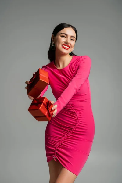 Веселая и очаровательная женщина с брюнетками и модными серёжками, улыбающаяся, стоя в пурпурном платье для вечеринок и держа упакованные подарочные коробки для отдыха на сером фоне — стоковое фото