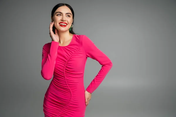 Элегантная и очаровательная женщина с брюнетками и модными серьгами, стоящая в пурпурном платье вечеринки, позируя с рукой на бедре и улыбаясь изолированы на сером фоне — стоковое фото