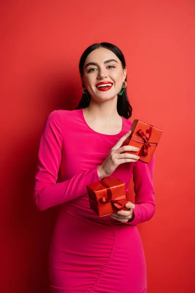 Joyful and charming woman with brunette hair and trendy earrings smiling while standing in magenta party dress and holding wrapped gift boxes for holiday on red background — Stock Photo