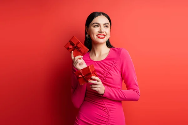 Fröhliche und elegante Frau mit brünetten Haaren, die lächelt, während sie im magentafarbenen Partykleid steht und verpackte Geschenkboxen für den Urlaub auf rotem Hintergrund hält — Stockfoto