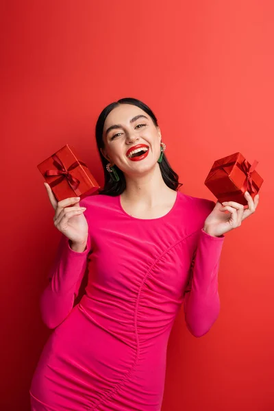 Donna eccitata e affascinante con capelli castani e orecchini alla moda sorridenti mentre in piedi in abito da festa magenta e in possesso di scatole regalo avvolte per le vacanze su sfondo rosso — Foto stock