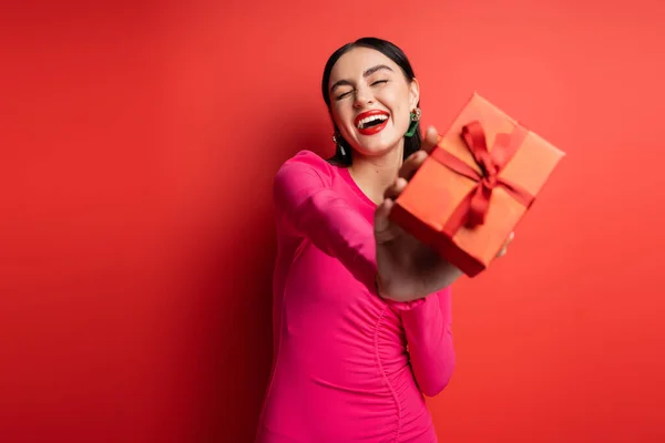 Aufgeregte Frau mit brünetten Haaren und trendigen Ohrringen lächelt, während sie im magentafarbenen Partykleid steht und eine verpackte Geschenkbox für den Urlaub auf rotem Hintergrund in der Hand hält — Stockfoto