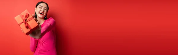 Mulher encantadora com cabelo morena e brincos na moda sorrindo enquanto estava de pé em vestido de festa magenta e segurando presente embrulhado para férias no fundo vermelho, banner — Fotografia de Stock