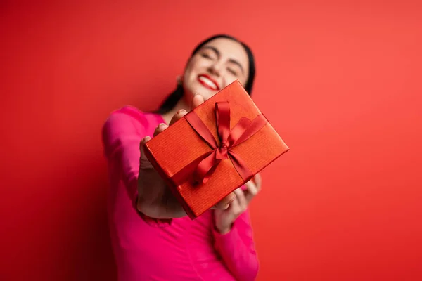 Happy blurred woman with brunette hair smiling while standing in magenta party dress and holding wrapped present with ribbon for holiday on red background — Stock Photo