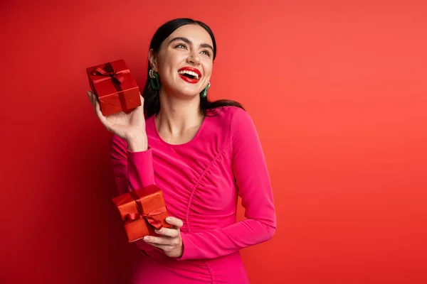 Mulher feliz com cabelo morena e brincos na moda sorrindo enquanto estava de pé em vestido de festa magenta e segurando caixas de presente embrulhado para férias no fundo vermelho — Fotografia de Stock