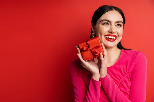 Gorgeous woman with brunette hair and trendy earrings smiling while standing in magenta party dress and holding wrapped present for holiday on red background — Stock Photo