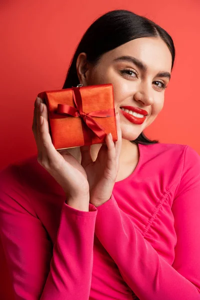 Portrait of gorgeous woman with brunette hair smiling while standing in magenta party dress and holding wrapped surprise gift for holiday on red background — Stock Photo