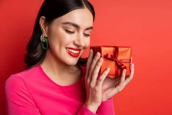Alluring woman with brunette hair and trendy earrings smiling while standing in magenta party dress and holding wrapped surprise gift for holiday on red background — Stock Photo