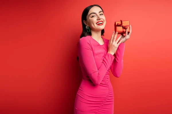 Блискуча жінка з брюнеткою волоссям і модними сережками посміхається, стоячи в вечірній сукні пурпурової вечірки і тримаючи загорнутий сюрприз подарунок на свято на червоному тлі — стокове фото