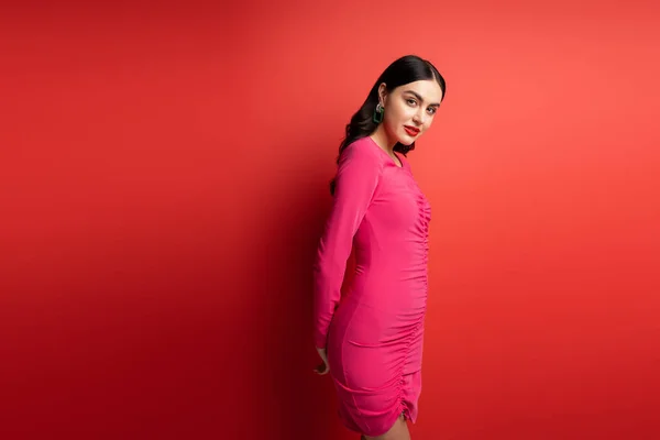 Glamorous woman with brunette hair and trendy earrings standing in magenta party dress while posing and looking at camera on red background — Stock Photo