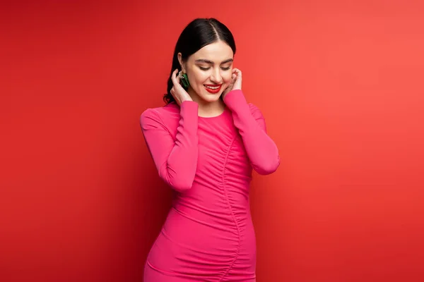 Glamorous woman with brunette hair and trendy earrings smiling while standing in magenta party dress while posing and looking down on red background — Stock Photo