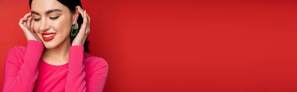 Mulher glamourosa com cabelo morena e brincos na moda sorrindo enquanto estava de pé em vestido de festa magenta e posando, olhando para o fundo vermelho, banner — Fotografia de Stock