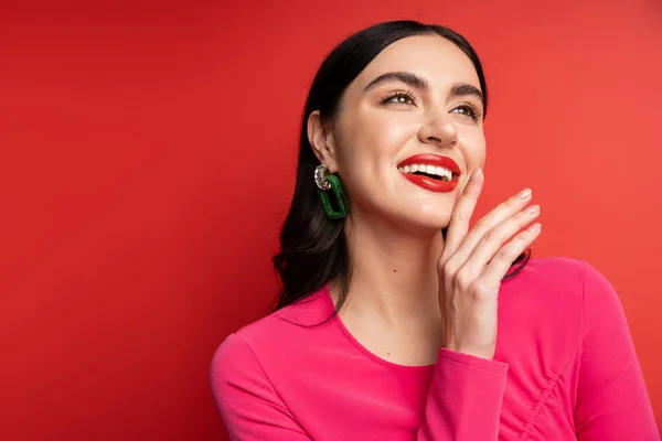 Portrait of pretty woman with brunette hair and trendy earrings smiling while standing in magenta party dress while posing with hand near face on red background — Stock Photo