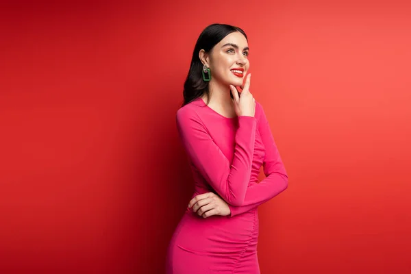 Красивая женщина с брюнетками и модными серёжками улыбаясь стоя в пурпурном платье вечеринки, позируя и глядя в сторону, думая на красном фоне — стоковое фото