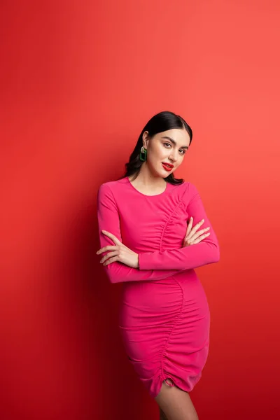Gorgeous woman with brunette hair and trendy earrings standing with folded arms in magenta party dress while posing and looking at camera on red background — Stock Photo