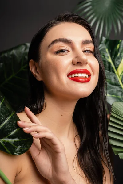 Очаровательная молодая женщина с брюнетками и красными губами улыбается, позируя вокруг тропических, влажных и зеленых пальмовых листьев с капельками дождя на них и глядя в камеру — стоковое фото