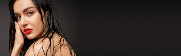 Молодая и очаровательная женщина с мокрыми и брюнетками, позирующая голыми плечами, глядя на камеру за душем, изолированную на сером фоне, баннер — стоковое фото