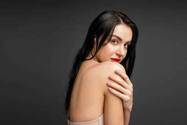 Portrait of young and graceful woman with wet brunette hair and red lips touching bare shoulder while looking at camera after shower isolated on grey background — Stock Photo