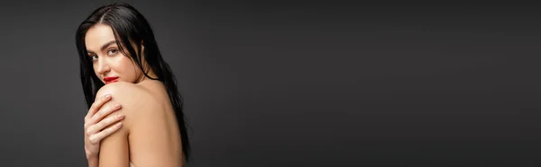 Porträt einer jungen und hübschen Frau mit nassen und brünetten Haaren, die die nackte Schulter berührt, während sie nach der Dusche isoliert auf grauem Hintergrund in die Kamera blickt, Banner — Stockfoto