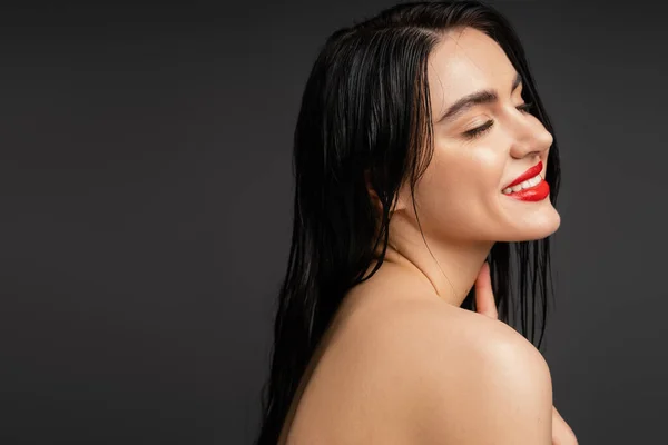 Retrato de mujer joven y feliz con el pelo moreno húmedo y labios rojos posando con hombro desnudo mientras sonríe con los ojos cerrados después de la ducha aislada sobre fondo gris - foto de stock