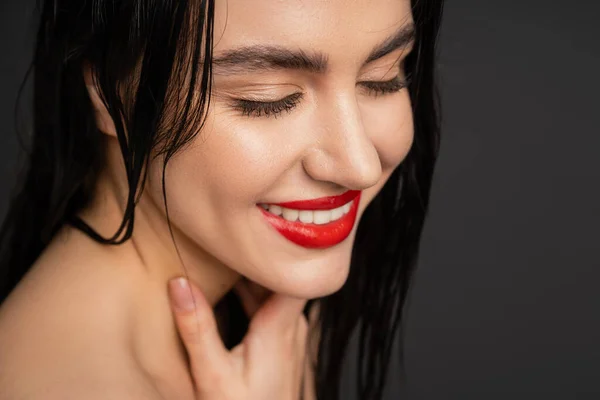 Close up vista de jovem e bonita mulher com cabelo morena molhada, lábios vermelhos posando com a mão no pescoço e sorrindo após o chuveiro isolado em fundo cinza — Fotografia de Stock