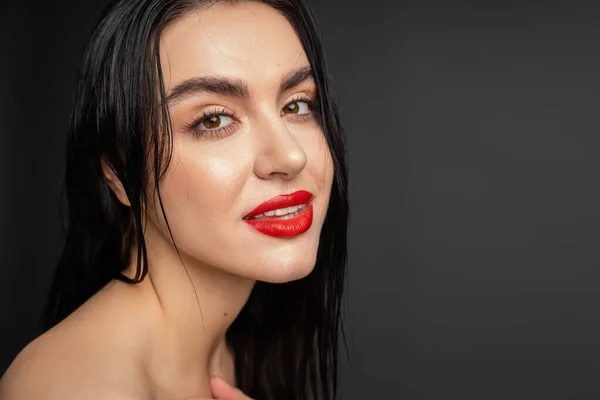 Porträt einer jungen und anmutigen Frau mit nassen brünetten Haaren und roten Lippen, die isoliert vor grauem Hintergrund in die Kamera blickt — Stockfoto