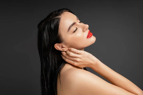 Gorgeous and graceful woman with wet brunette hair and red lips touching neck while posing with bare shoulders after shower isolated on grey background — Stock Photo