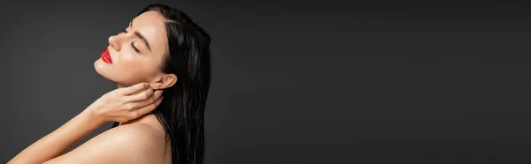 Красивая и молодая женщина с мокрыми волосами брюнетки и красными губами касаясь шеи, позируя с голыми плечами после душа изолированы на сером фоне, баннер — стоковое фото