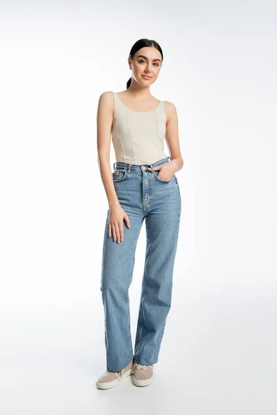Full length of charming young brunette woman standing in blue denim jeans and tank top while posing with hand in pocket and looking at camera on white background — Stock Photo