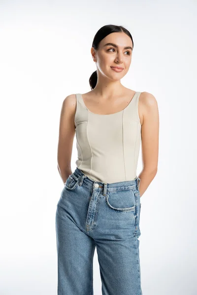 Charming and young brunette woman standing in blue denim jeans and tank top while smiling, posing and looking away on white background — Stock Photo