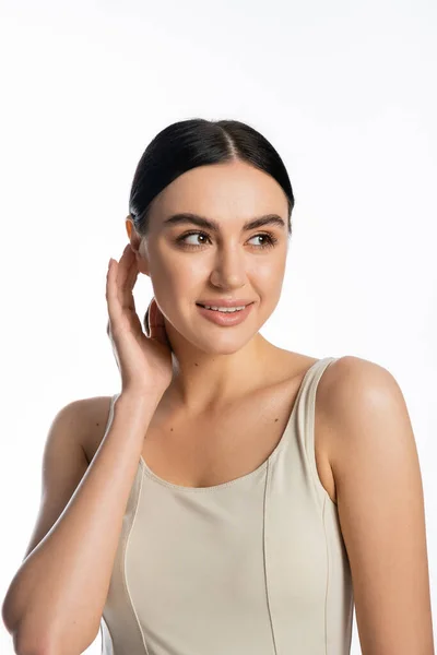 Portrait of charming young woman with flawless and natural makeup standing in tank top while adjusting brunette hair and looking away isolated on white background — Stock Photo