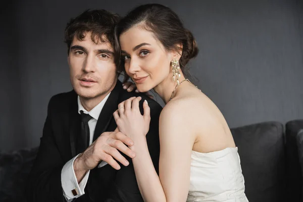 Portrait of charming and young brunette bride in luxurious earrings with pearls and white wedding dress hugging positive groom in black suit in hotel room — Stock Photo