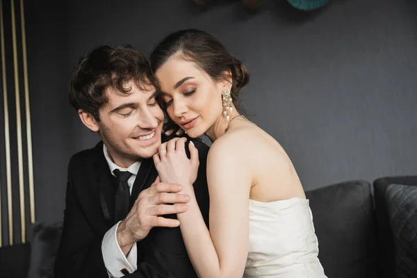 Portrait of young brunette bride in luxurious earrings with pearls and white wedding dress sitting with closed eyes and hugging happy groom in black suit in hotel room — Stock Photo