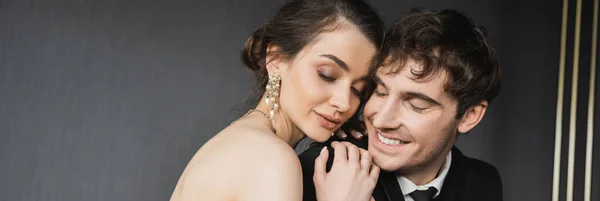 Portrait of young brunette bride in luxurious earrings with pearls sitting with closed eyes and hugging happy groom in black suit in hotel room, banner — Stock Photo