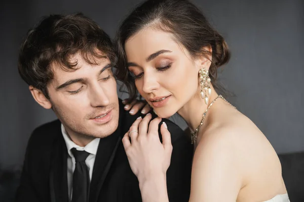 Ritratto di bella giovane sposa in lussuosi orecchini con perle e collana seduta ad occhi chiusi e abbracciare felice sposo in abito nero in camera d'albergo — Foto stock